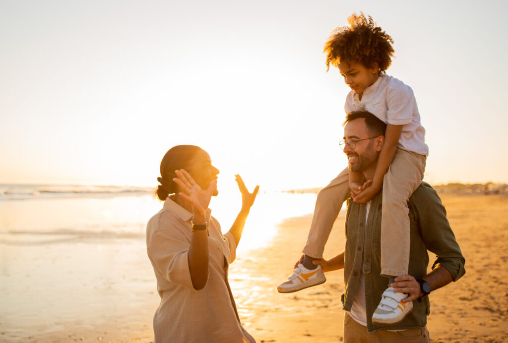 Sand, sea and smiles. Happy young diverse family of three spending free time outdoors, walking and having fun on seaside in the evening, father holding son on shoulders